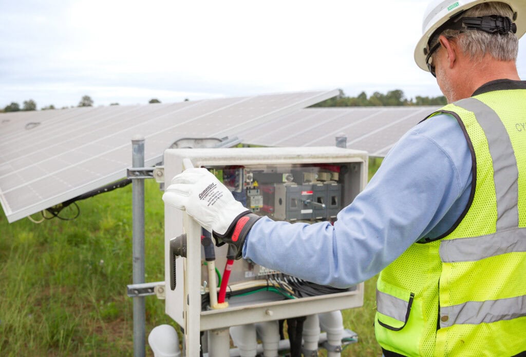 A man in safety gear opens an electrical box next to a solar panel in a green field