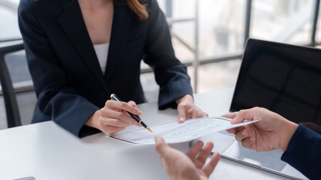 Two professionals sitting at a table with one person handing a paper to the other person who is also holding a pen.