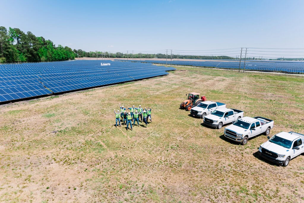 From an aerial view a group of people wave as they stand in a large field between rows of solar panels and work trucks as well as a tractor.