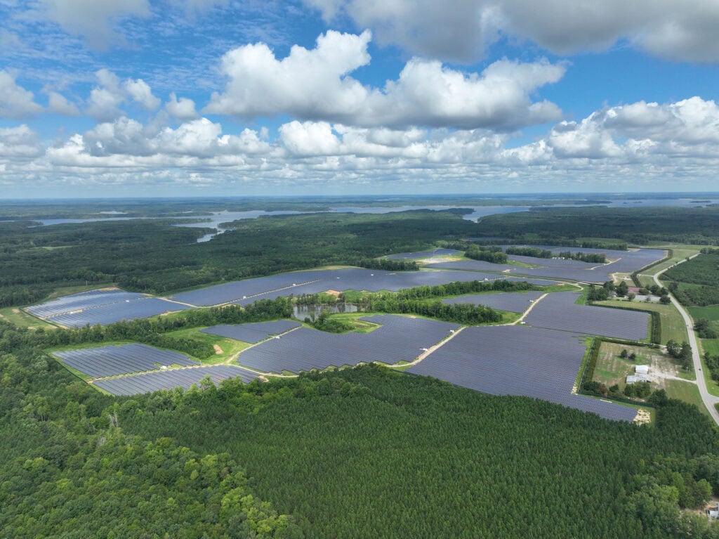 A solar farm surrounded by green trees is seen in an aerial view with a blue sky and far off image of a lake in the background.